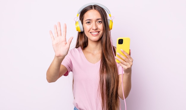 Mulher jovem e bonita sorrindo e parecendo amigável, mostrando o número cinco. fones de ouvido e smartphone