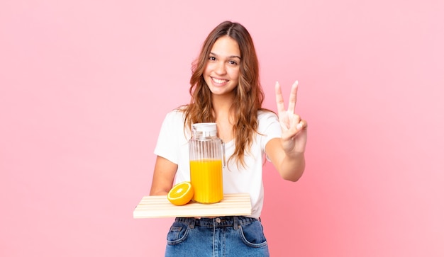 Mulher jovem e bonita sorrindo e parecendo amigável, mostrando o número dois e segurando uma bandeja com um suco de laranja