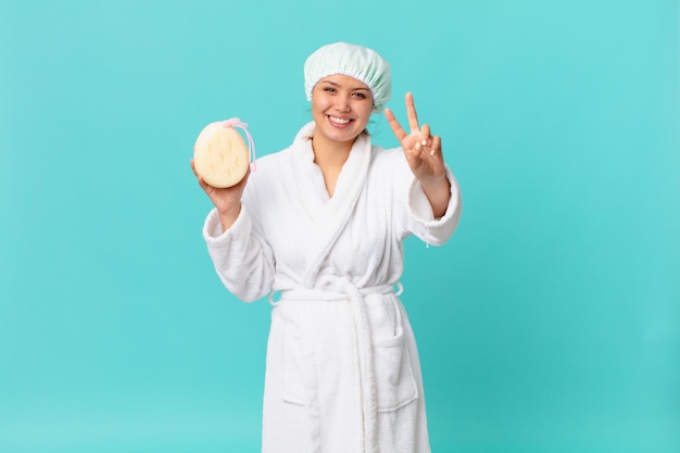 Mulher jovem e bonita sorrindo e parecendo amigável, mostrando o número dois e usando roupão de banho após o banho