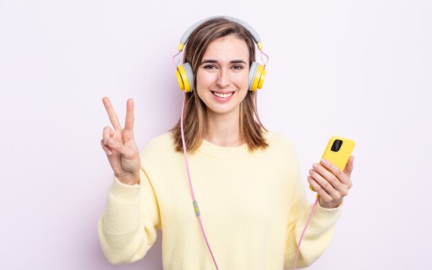 Mulher jovem e bonita sorrindo e parecendo amigável, mostrando o número dois. fones de ouvido e conceito de telefone