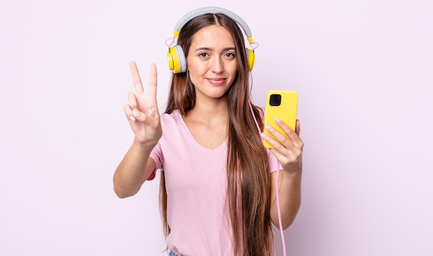 Mulher jovem e bonita sorrindo e parecendo amigável, mostrando o número dois. fones de ouvido e smartphone