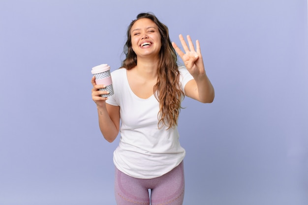 Mulher jovem e bonita sorrindo e parecendo amigável, mostrando o número quatro e segurando um café