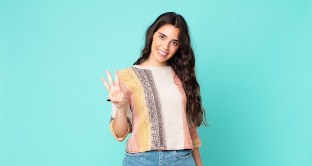 Mulher jovem e bonita sorrindo e parecendo amigável, mostrando o número três ou o terceiro com a mão para a frente, em contagem regressiva