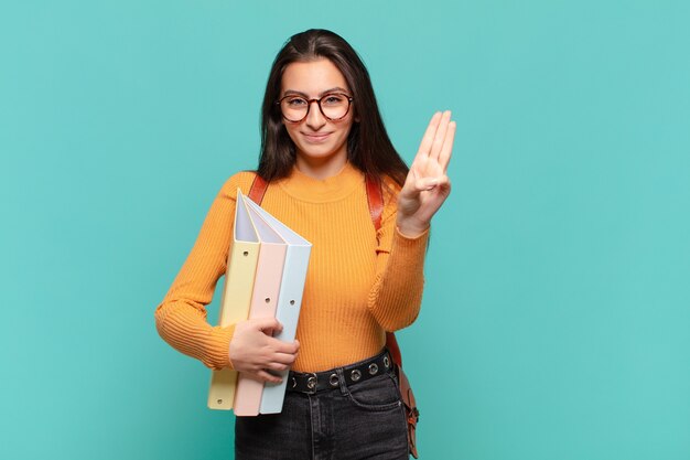 Mulher jovem e bonita sorrindo e parecendo amigável, mostrando o número três ou terceiro com a mão para a frente, em contagem regressiva. conceito de estudante