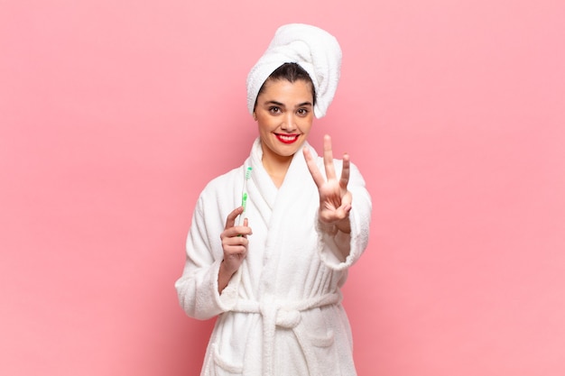 Mulher jovem e bonita sorrindo e parecendo amigável, mostrando o número três ou terceiro com a mão para a frente, em contagem regressiva. conceito de roupão e escova de dentes