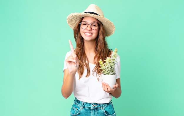 Mulher jovem e bonita sorrindo e parecendo amigável, mostrando o número um com um chapéu de palha e segurando um cacto