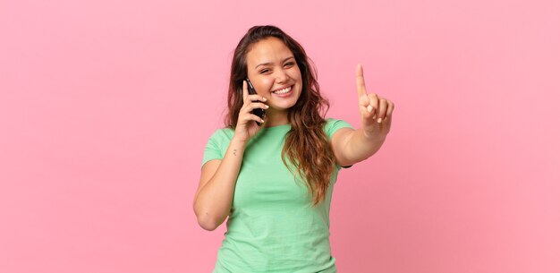 Mulher jovem e bonita sorrindo e parecendo amigável, mostrando o número um e segurando um telefone inteligente