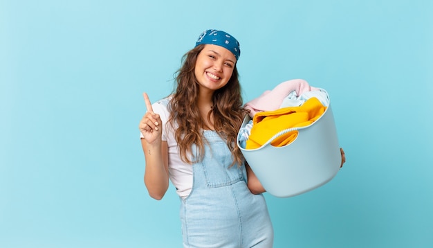 Mulher jovem e bonita sorrindo e parecendo amigável, mostrando o número um e segurando uma cesta de roupas para lavar