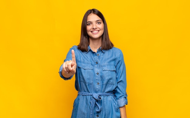 Mulher jovem e bonita sorrindo e parecendo amigável, mostrando o número um ou primeiro com a mão para a frente, em contagem regressiva