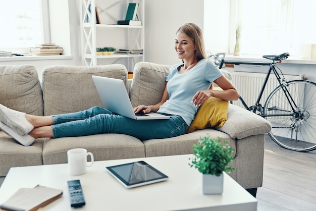 Mulher jovem e bonita sorrindo e usando o laptop enquanto descansa no sofá em casa