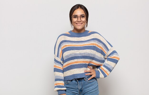 Foto mulher jovem e bonita sorrindo feliz com uma mão no quadril e com uma atitude confiante, positiva, orgulhosa e amigável