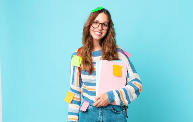 Mulher jovem e bonita sorrindo feliz com uma mão no quadril e confiante com uma bolsa e segurando livros