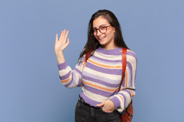 Mulher jovem e bonita sorrindo feliz e alegremente, acenando com a mão, dando as boas-vindas e cumprimentando-o ou dizendo adeus. conceito de estudante