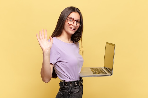 Mulher jovem e bonita sorrindo feliz e alegremente, acenando com a mão, dando as boas-vindas e cumprimentando-o ou dizendo adeus. conceito de laptop