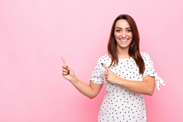 Mulher jovem e bonita sorrindo feliz e apontando para o lado e para cima com as duas mãos mostrando o objeto no espaço da cópia contra a parede rosa