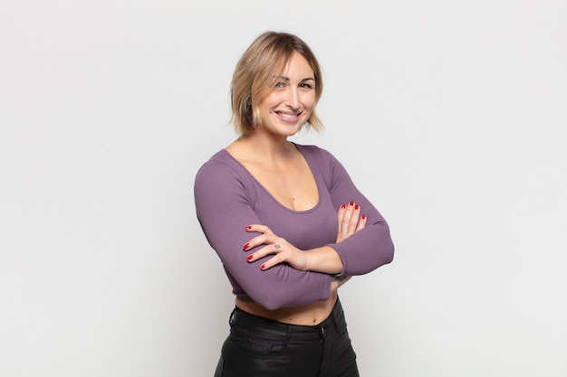 Mulher jovem e bonita sorrindo para a câmera com os braços cruzados e uma expressão feliz, confiante e satisfeita, vista lateral
