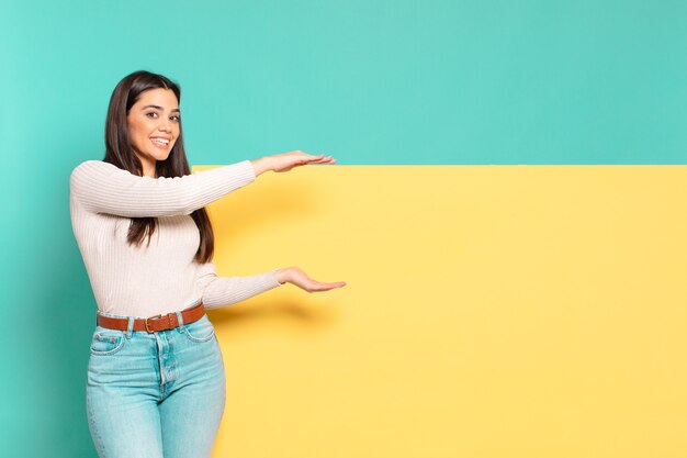 Mulher jovem e bonita sorrindo, sentindo-se feliz, positiva e satisfeita, segurando ou mostrando o objeto ou conceito no espaço da cópia. copie o espaço para colocar o seu conceito