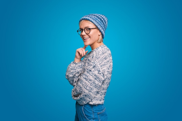 Mulher jovem e bonita sorrindo suavemente, mantendo a mão sob o queixo olhando para a câmera com expressão sonhadora usando chapéu de malha isolado sobre fundo azul