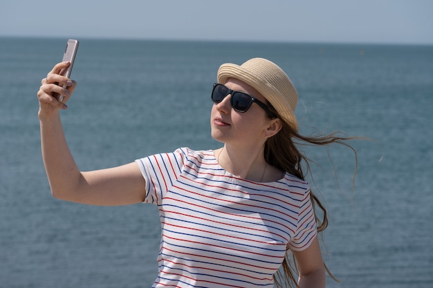 Mulher jovem e bonita tira uma foto de si mesma ao telefone no contexto do porto marítimo azul ...