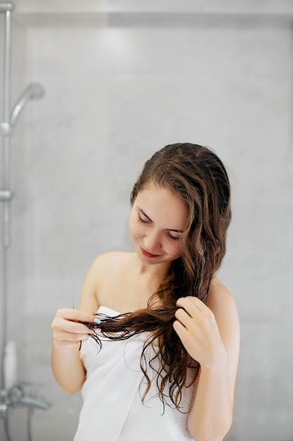 Mulher jovem e bonita tocando seu cabelo molhado com as mãos e sorrindo em pé na frente do espelho no banheiro. Conceito de cuidados com a pele do cabelo