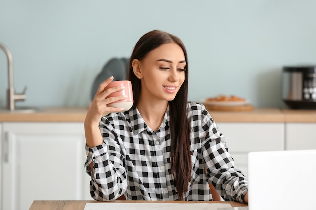 Mulher jovem e bonita tomando chá e usando o laptop na cozinha