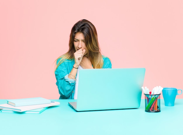 Mulher jovem e bonita trabalhando com um laptop, sentindo-se doente com sintomas de dor de garganta e gripe, tosse com a boca coberta