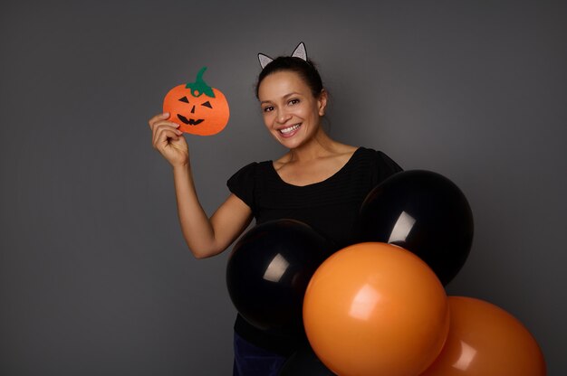 Mulher jovem e bonita usando aro com orelhas de gato, vestida em traje preto, segurando bolas de ar coloridas pretas e laranja e mostra uma abóbora artesanal cortada em feltro, sorri olhando para a câmera. Conceito de halloween