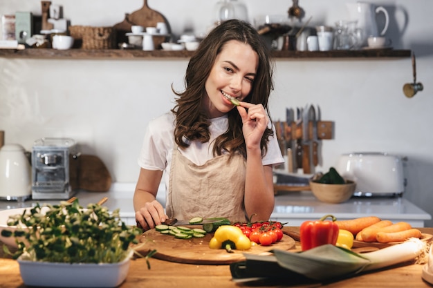 Mulher jovem e bonita usando avental cozinhando salada saudável na cozinha em casa, cortando vegetais