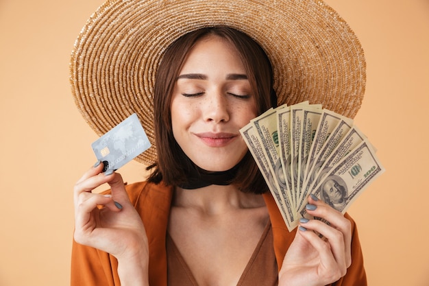 Foto mulher jovem e bonita usando chapéu de palha e roupa de verão em pé, isolada na parede bege, segurando um cartão de crédito palstic, mostrando notas de dinheiro