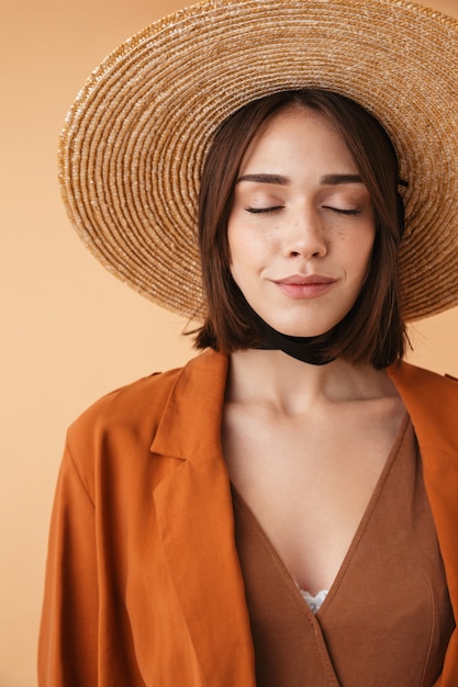 Mulher jovem e bonita usando chapéu de palha e roupa de verão em pé, isolada sobre uma parede bege, posando