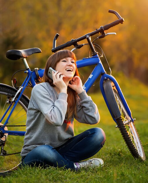 Mulher jovem e bonita usando telefone celular, sentado ao lado de bicicleta ao pôr do sol