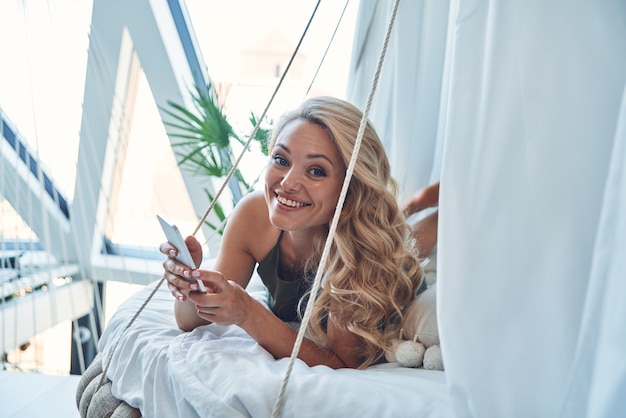 Mulher jovem e bonita usando telefone inteligente e sorrindo enquanto relaxa na cama em casa