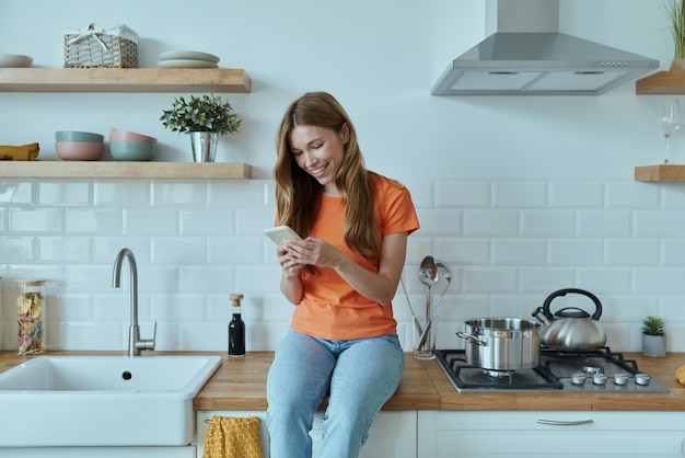 Mulher jovem e bonita usando telefone inteligente enquanto está sentado no balcão da cozinha