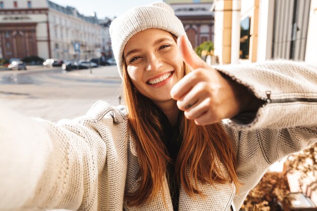 Mulher jovem e bonita vestida com casaco outono e chapéu caminhando ao ar livre, tirando uma selfie