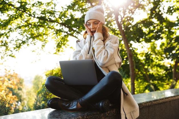 Mulher jovem e bonita vestida com um casaco e chapéu outono sentada ao ar livre, usando um computador laptop, comemorando o sucesso