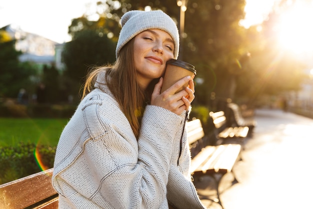 Mulher jovem e bonita vestida com um casaco outono e um chapéu sentada em um banco ao ar livre, segurando uma xícara de café quente