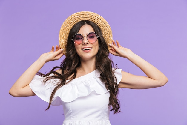 Mulher jovem e bonita vestindo roupa de verão e óculos escuros em pé isolado sobre a parede violeta, posando
