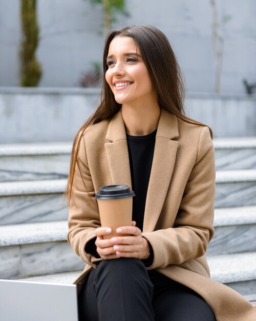 Mulher jovem e bonita vestindo um casaco de outono, sentada em um banco, bebendo café para viagem