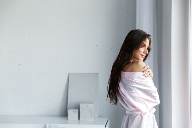 Mulher jovem e bonita vestindo um manto em pé na janela do quarto