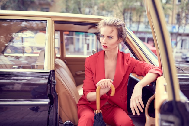 Mulher jovem e bonita vestindo uma fantasia vermelha, segurando um guarda-chuva enquanto está sentado em um carro retrô