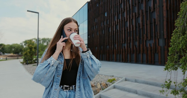 Mulher jovem e bonita vestindo uma jaqueta jeans e usando smartphone moderno enquanto caminhava no intervalo na cidade mulher falando no telefone com amigos regozijando-se sorrindo rindo e bebendo café