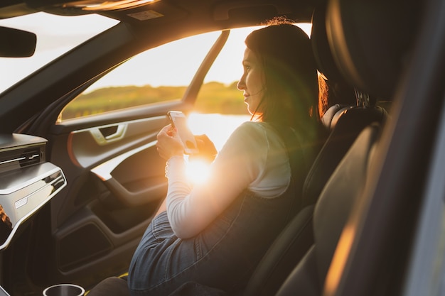 Mulher jovem e bonita viaja nas estradas de carro estacionado na beira da estrada e usa smartphone