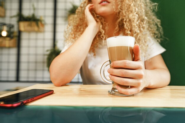 Mulher jovem e cacheada sentada à mesa na cafeteria e relaxando com uma xícara de café, retrato