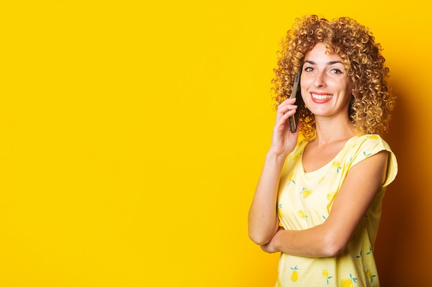 Mulher jovem e cacheada sorrindo e falando ao telefone em uma superfície amarela