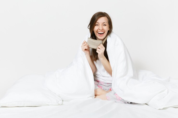 Foto mulher jovem e cansada divertida sentada na cama com máscara de dormir, lençol, travesseiro, embrulhado em cobertor isolado na parede branca