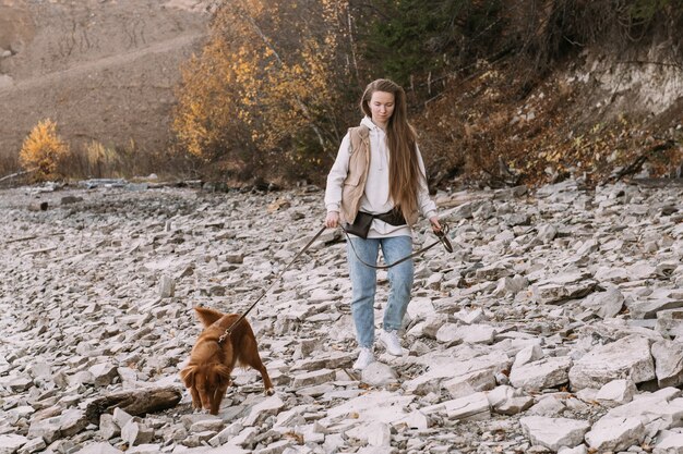 Mulher jovem e cão retriever caminham na margem do rio na temporada de outono