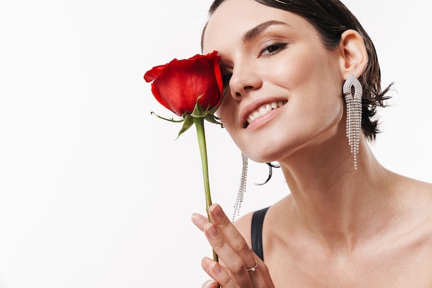 Mulher jovem e charmosa e glamorosa sorrindo e segurando uma flor isolada na parede branca