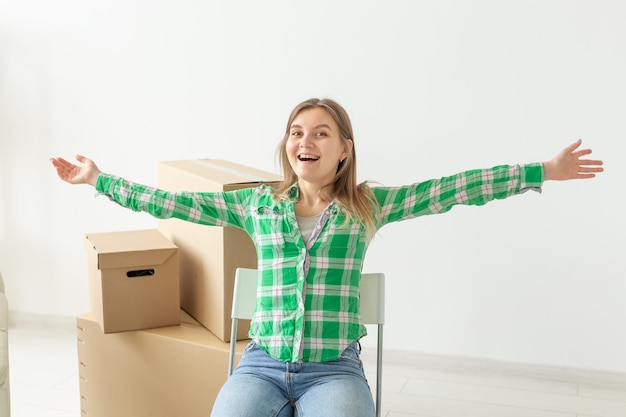 Mulher jovem e charmosa em roupas casuais se regozija sentada em uma cadeira entre as caixas de coisas em sua sala de estar em um novo apartamento durante a mudança. conceito de nova habitação e inauguração de casa. copyspace