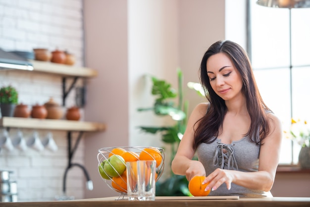 Mulher jovem e desportiva está cortando laranja fresca para suco de frutas na cozinha. tiro horizontal em ambientes fechados.