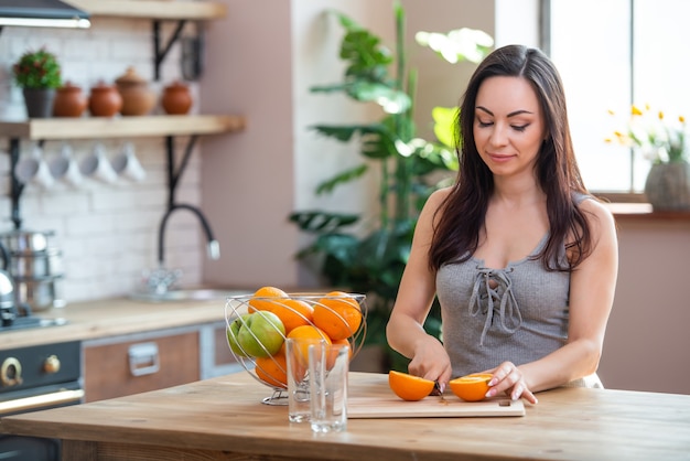 Mulher jovem e desportiva está cortando laranja fresca para suco de frutas na cozinha. Tiro horizontal em ambientes fechados.